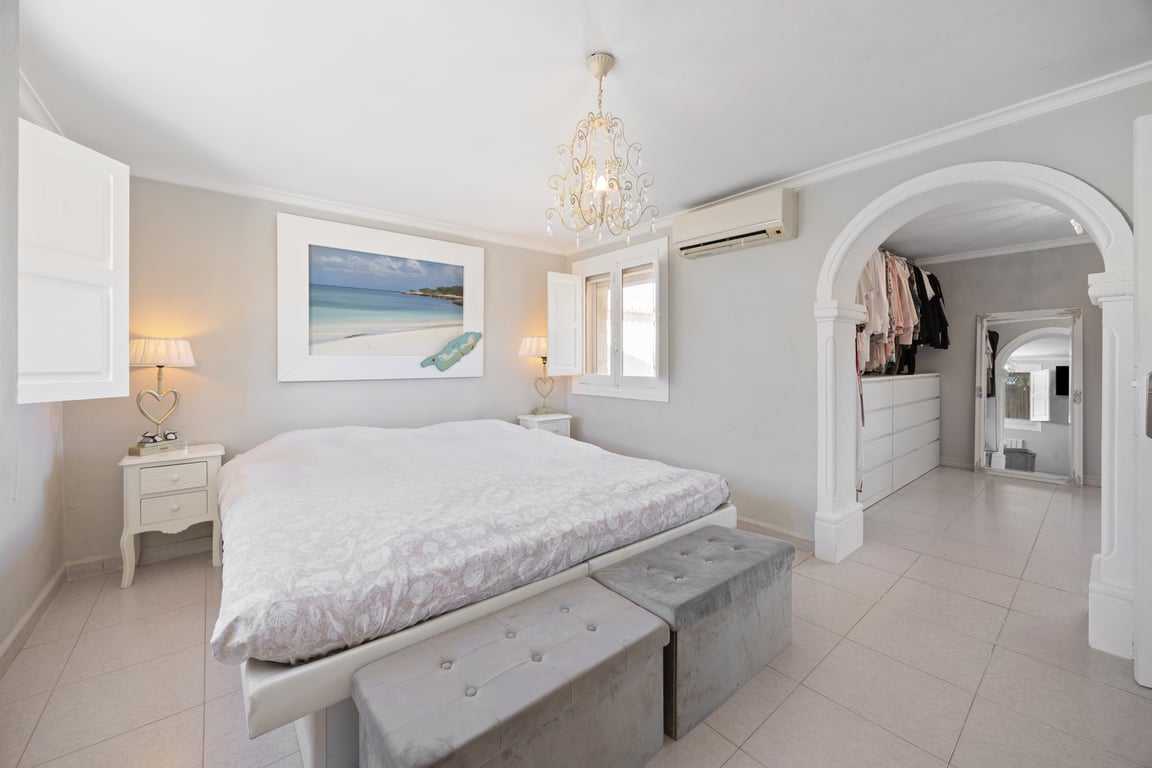 Hoofdslaapkamer met inloopkast achter een decoratieve boog, tegelvloer, airconditioning en ramen met witte houten binnenluiken.