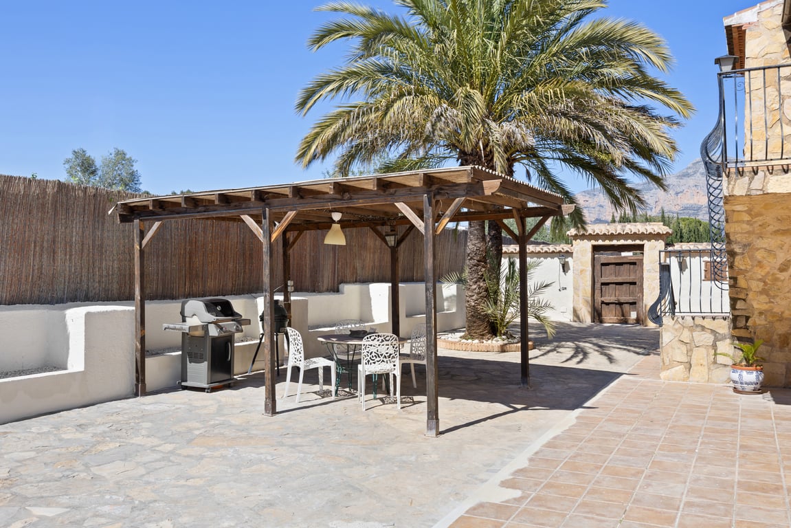 Houten pergola met een eethoek buiten en barbecue, gelegen op een stenen terras met een volwassen palmboom en uitzicht op de bergen.