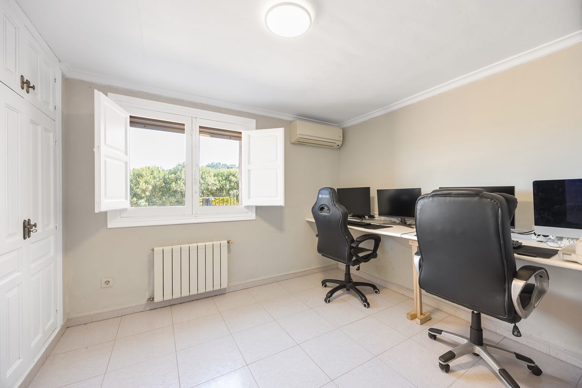 Licht kantoor met inbouwkasten, airconditioning en radiator. Het raam met binnenluiken zorgt voor natuurlijk licht en uitzicht op de omliggende tuin.