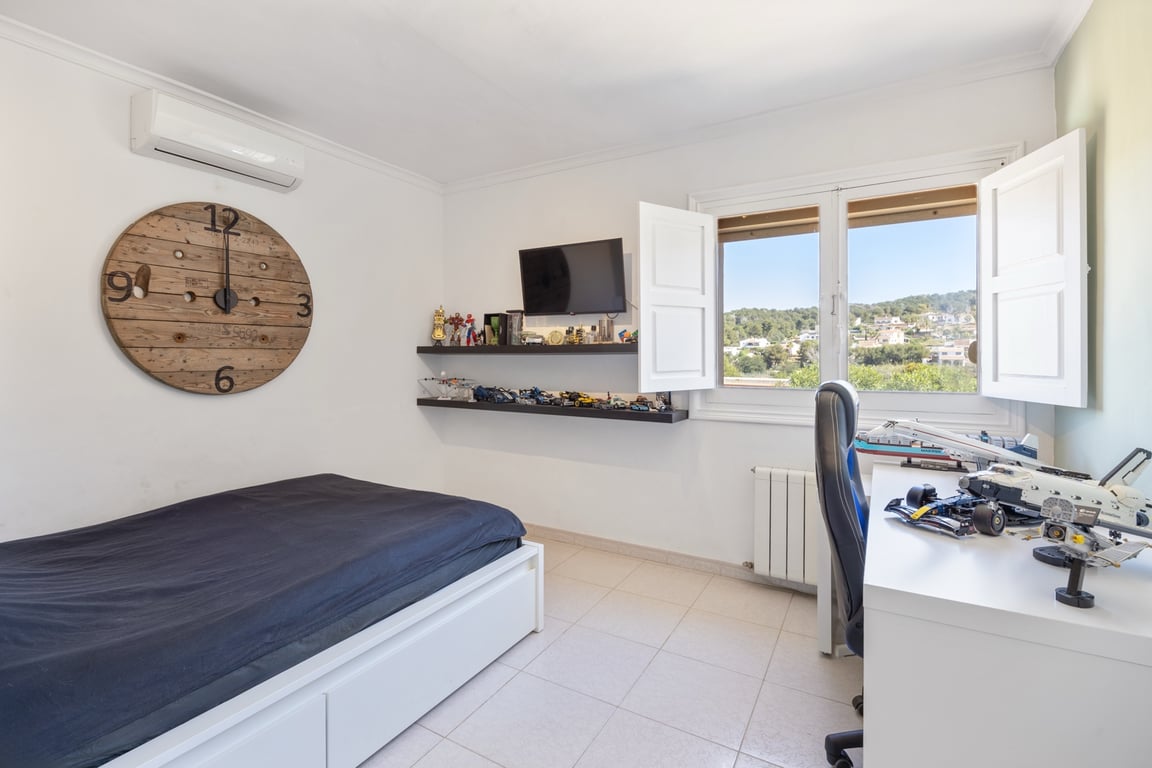 Lichte slaapkamer met airconditioning, een bureau en een raam met witte luiken dat uitzicht biedt op de omliggende groene heuvels.