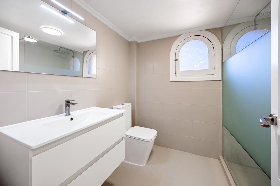 Moderne badkamer met wit wastafelmeubel, inloopdouche met glazen wand en een karakteristiek boograam. Neutrale betegeling en heldere LED-verlichting boven de spiegel.