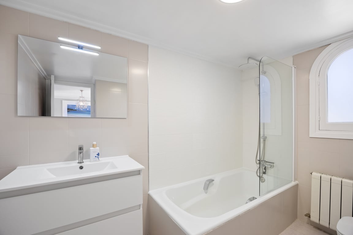 Moderne badkamer met wit wastafelmeubel, verlichte spiegel en ligbad met glazen douchescherm. Een boogvenster biedt natuurlijk licht bij de neutrale betegeling.