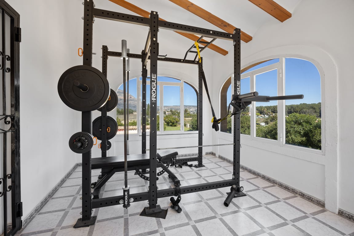 Privé-fitnessruimte uitgerust met professionele Rogue-apparatuur, gelegen in een lichte ruimte met houten balken en vrij uitzicht op de mediterrane omgeving.