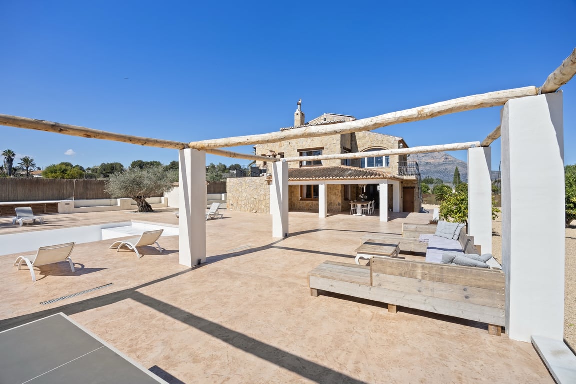 Ruim buitenterras met een houten balkenpergola, loungehoek en uitzicht op de stenen gevel van de villa en de berg Montgó.