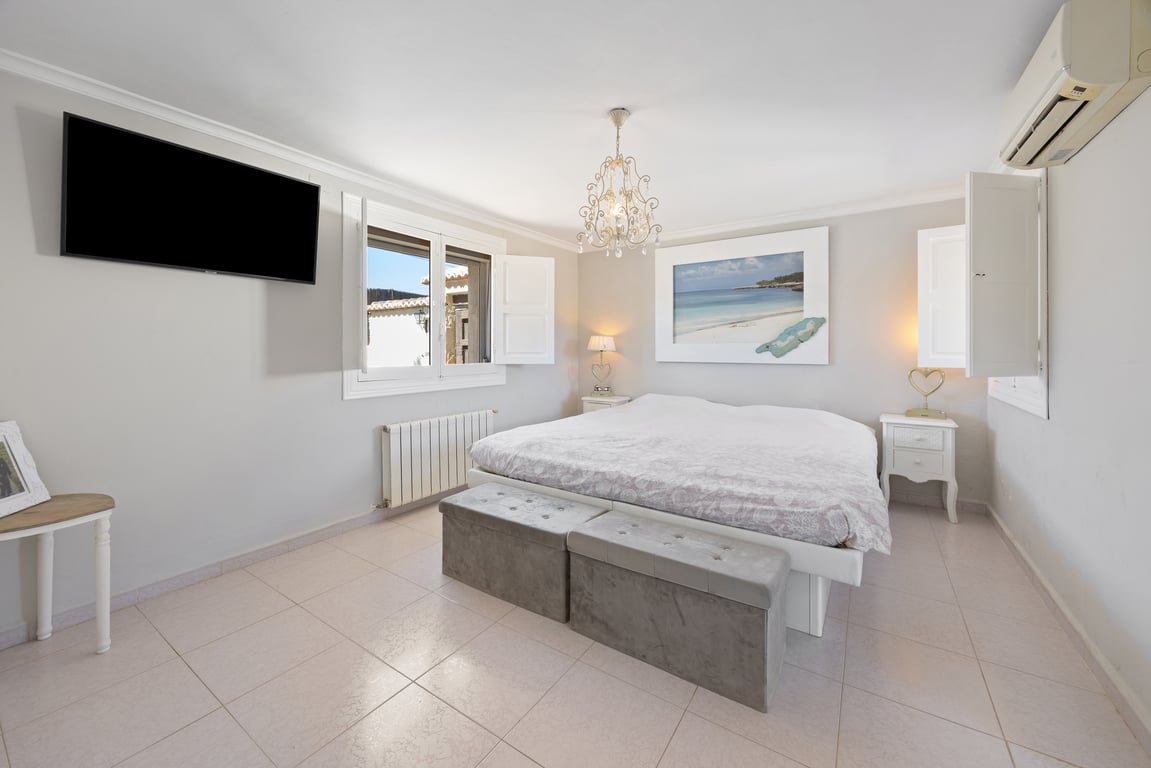 Ruime slaapkamer met witte luiken, een wand-tv en airconditioning. De kamer is voorzien van een groot bed met gestoffeerde bankjes en een lichte tegelvloer.