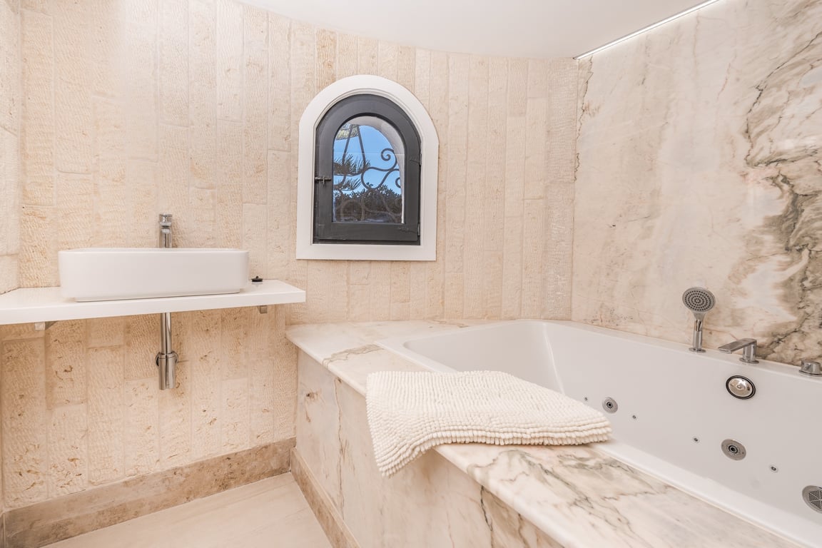 Badkamer met een in marmer afgewerkt bubbelbad, opzetwastafel en natuurstenen wanden voorzien van een karakteristiek rondboogvenster.