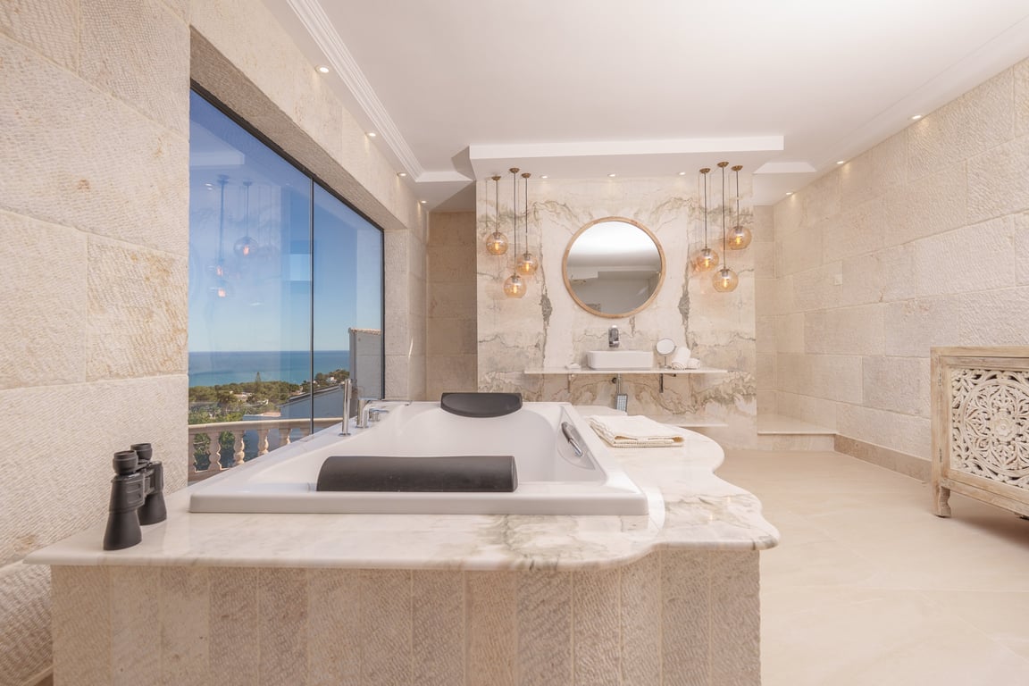 Badkamer van de master suite met kalkstenen muren, een in marmer ingebouwd ligbad met zeezicht en een designwastafel met decoratieve hangverlichting.