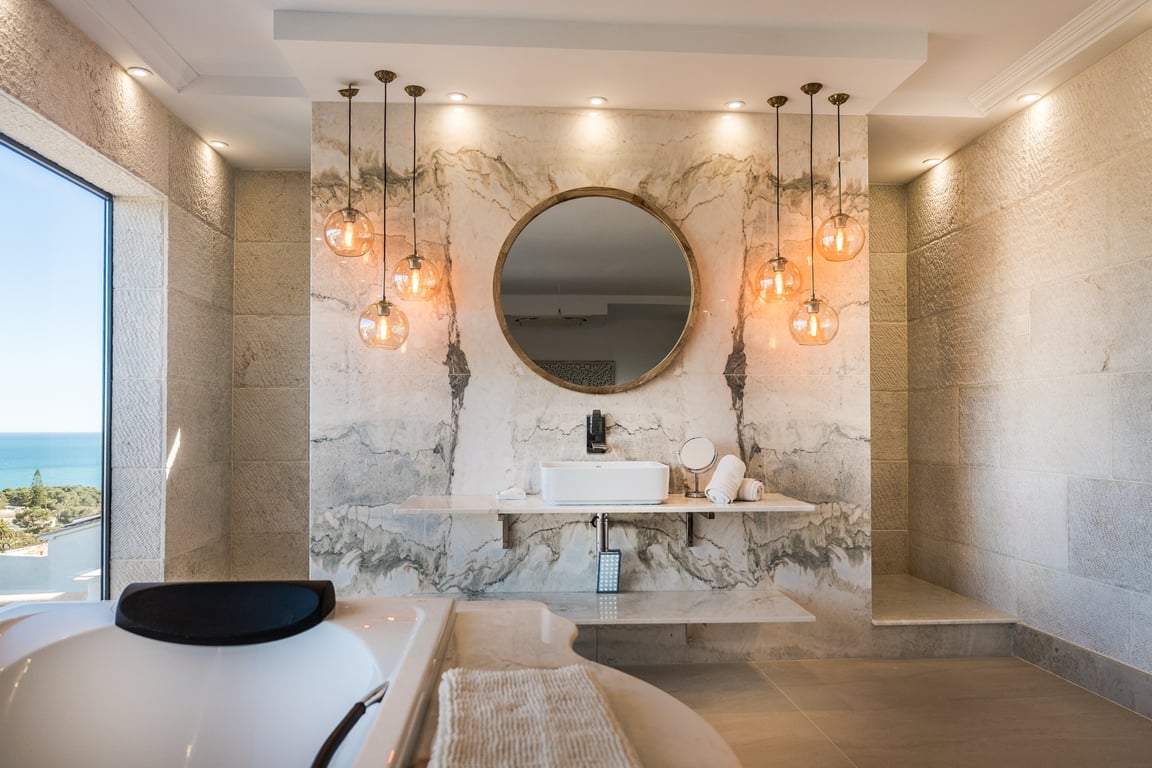 Badkamer van de master suite met natuurstenen wanden, geaderd marmeren wastafelblad, bolvormige hanglampen en weids uitzicht op zee.