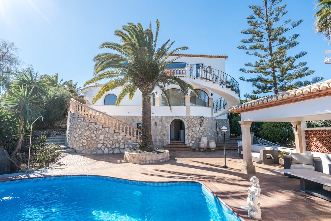 Buitenaanzicht van de villa met zwembad, natuurstenen gevel, sierlijke wenteltrappen en een mediterrane tuin met palmbomen in de zon van Jávea.