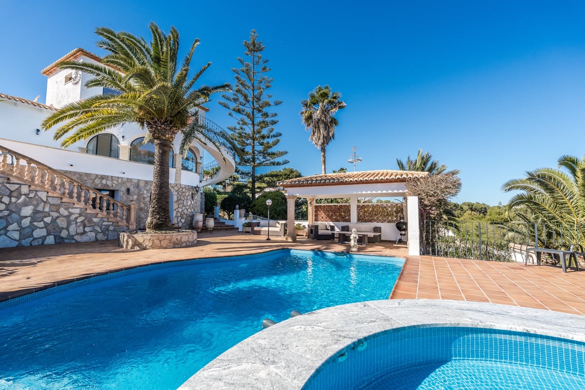Buitenruimte met zwembad en jacuzzi, omgeven door een terracotta terras, palmbomen en een villa met natuurstenen gevel, klassieke trap en overdekt loungeterras.