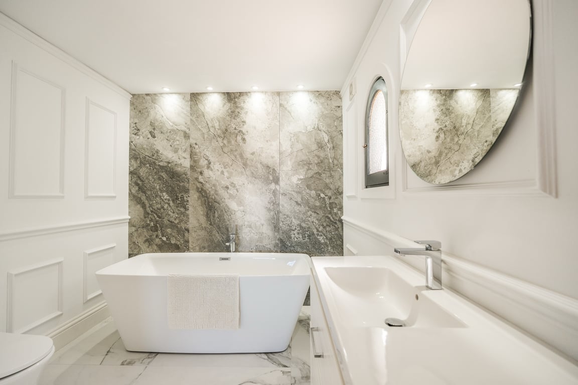 Elegante badkamer met vrijstaand bad, marmeren accentmuur met inbouwspots en klassieke witte wandpanelen voor een tijdloze uitstraling.