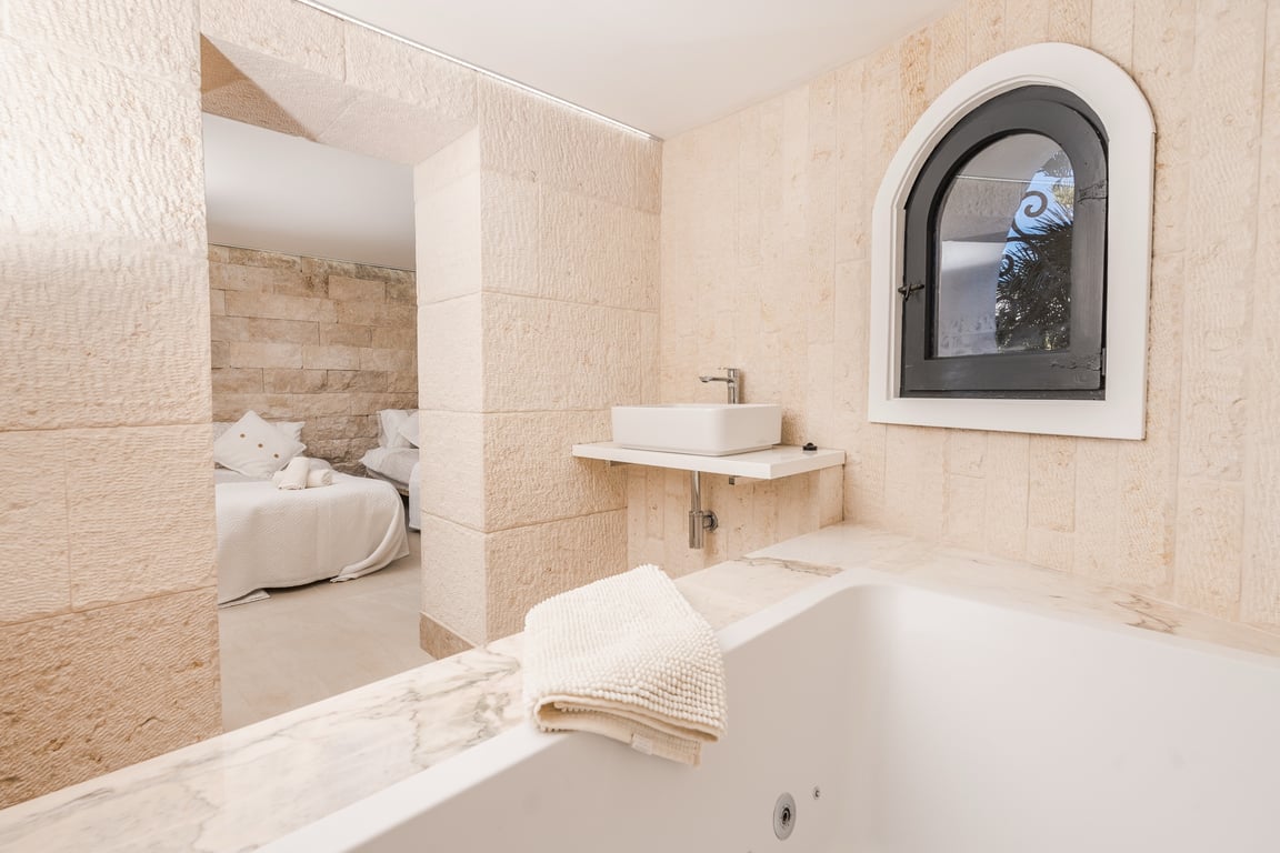 En-suite badkamer met natuurstenen muren, een ligbad met marmeren rand en een moderne wastafel, met direct zicht op de aangrenzende slaapkamer via een stenen boog.