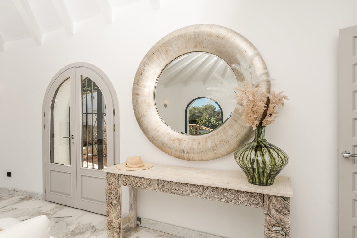 Hal met marmeren vloer, een bewerkte houten console en een grote ronde spiegel. Witte balkenplafonds en boogdeuren benadrukken het verfijnde mediterrane karakter.