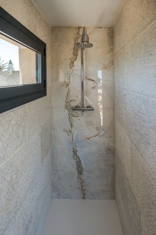 Inloopdouche met een achterwand van geaderd marmer en zijwanden van getextureerde kalksteen. Voorzien van chromen kranen en een raam voor natuurlijke ventilatie.