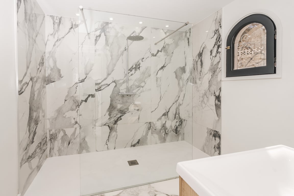 Inloopdouche met marmerlook wandtegels, glazen wand en een authentiek rondboogvenster met zwart kozijn en smeedijzeren rooster voor een unieke uitstraling.