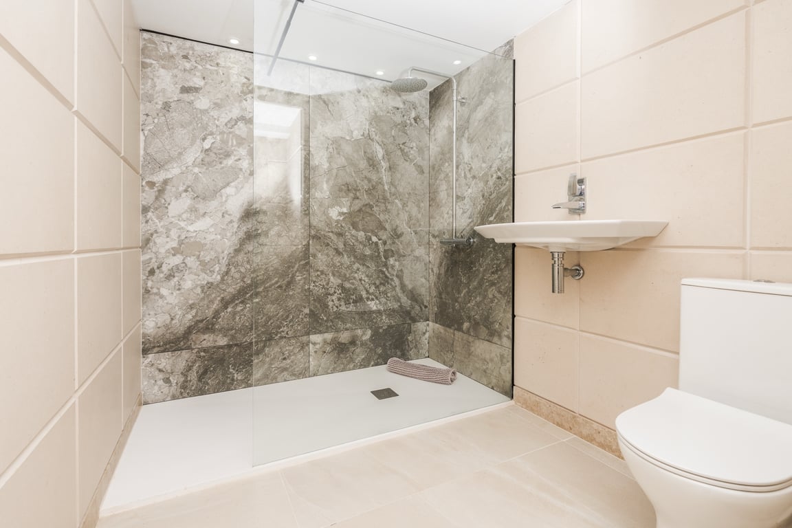 Moderne badkamer met inloopdouche, glazen wand en achterwand met marmerlook. Voorzien van wit minimalistisch sanitair en crème wandtegels.