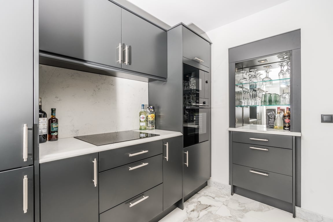 Moderne keuken met antracietgrijze kasten, wit marmeren werkbladen en geïntegreerde apparatuur. Inclusief een bar met verlichte vitrinekast en een stijlvolle marmeren vloer.