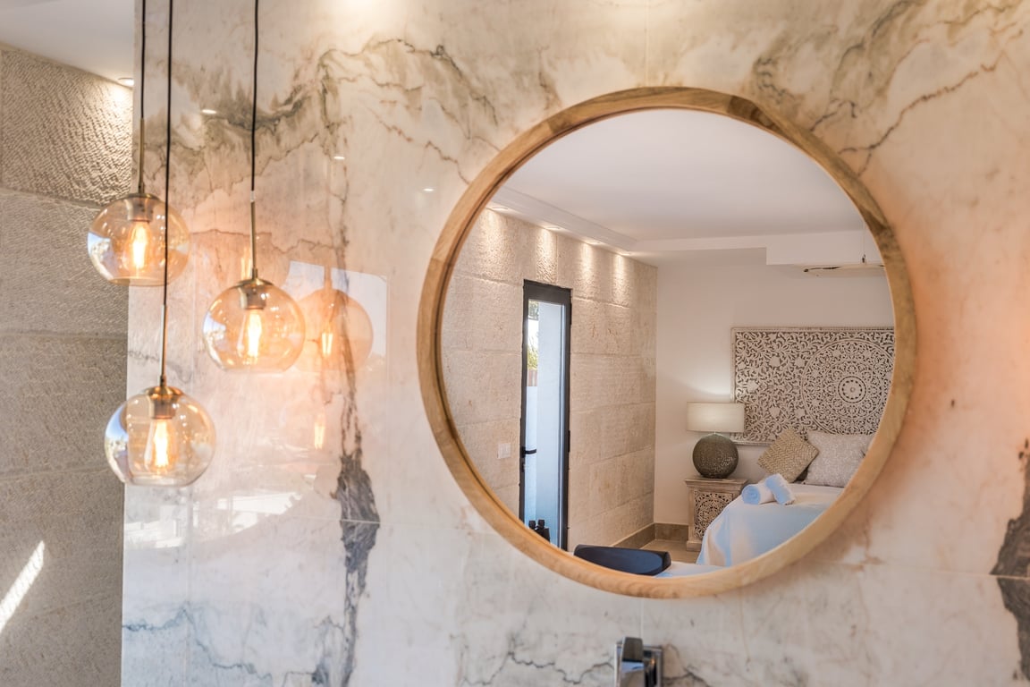 Ronde spiegel met houten lijst op een geaderde marmeren wand, voorzien van glazen hanglampen en reflectie van de slaapkamer met kalkstenen muren.