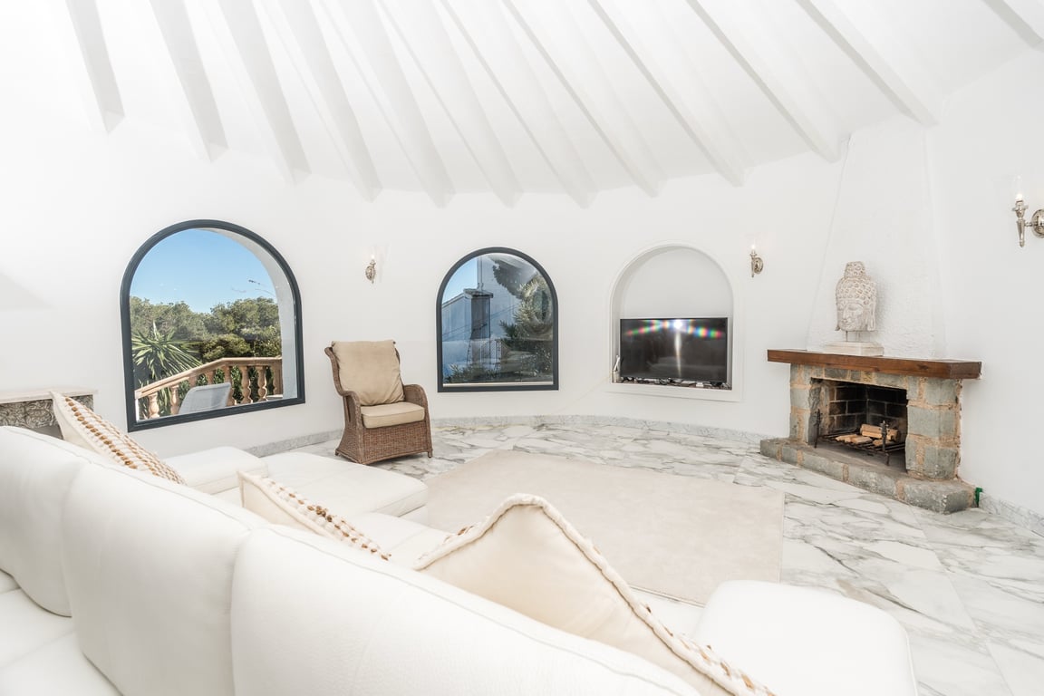 Ronde woonkamer met wit balkenplafond, marmeren vloer en een stenen open haard. Boogramen zorgen voor natuurlijk licht en uitzicht op de mediterrane omgeving.