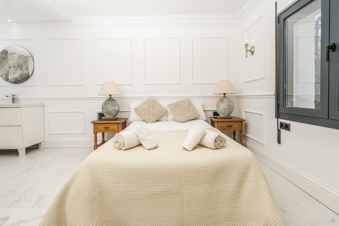Slaapkamer met witte wandpanelen en marmeren vloer. Inclusief een geïntegreerd wastafelmeubel met ronde spiegel en klassieke houten nachtkastjes.