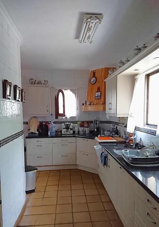 Aparte keuken voorzien van witte kasten, donkere werkbladen en een terracotta tegelvloer. Inclusief een houten afzuigkap en natuurlijke lichtinval via het raam.