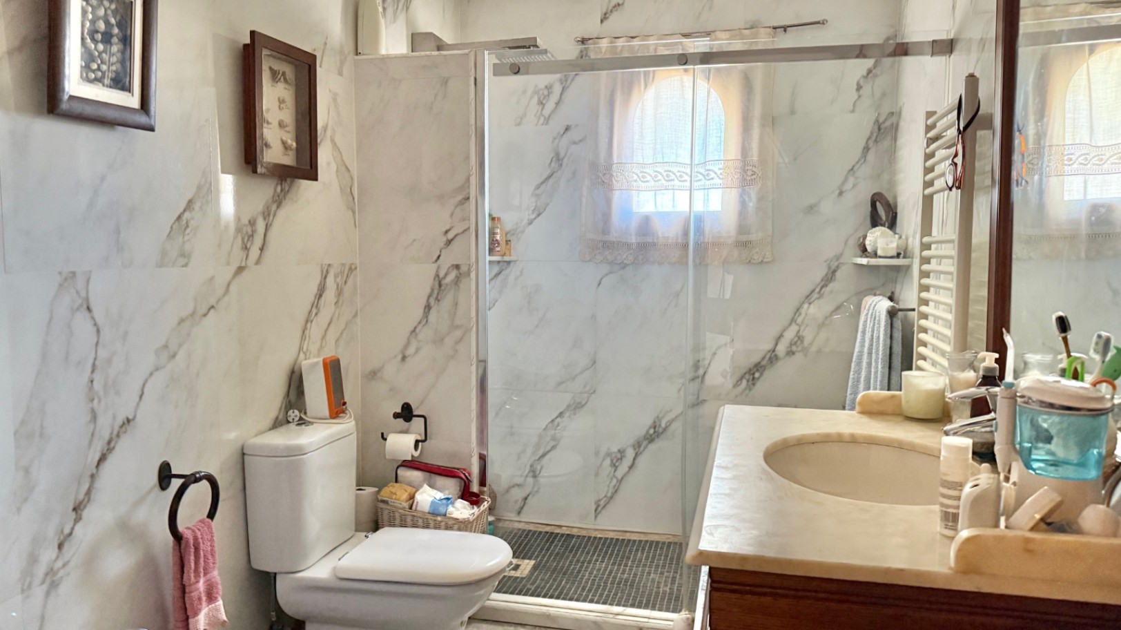 Badkamer met wandtegels in marmerlook, inloopdouche met glazen wand en een marmeren wastafelblad. Voorzien van een handdoekradiator en een klein boogvenster.