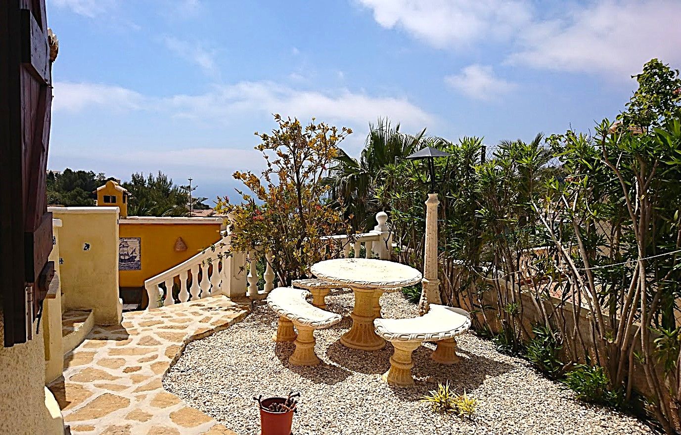 Buitenzitplaats met een bewerkte stenen tafel en banken op een grindterras, omringd door mediterrane beplanting en uitzicht op zee.