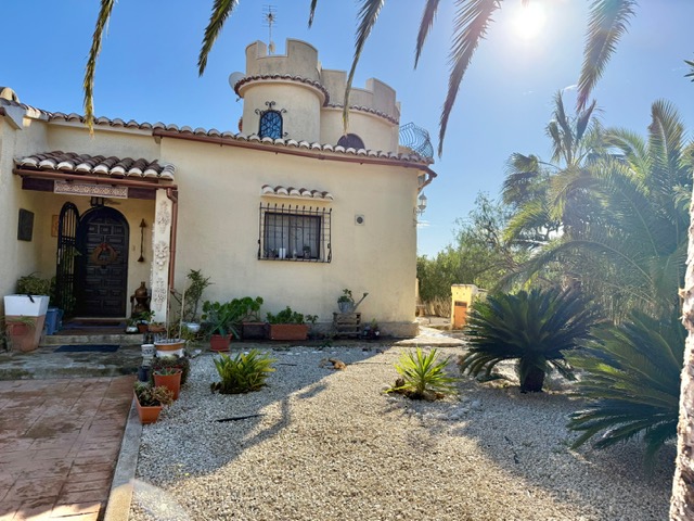Hoofdfaçade van de villa met de kenmerkende ronde toren, overdekte entree en een mediterrane grindtuin met volwassen palmbomen.