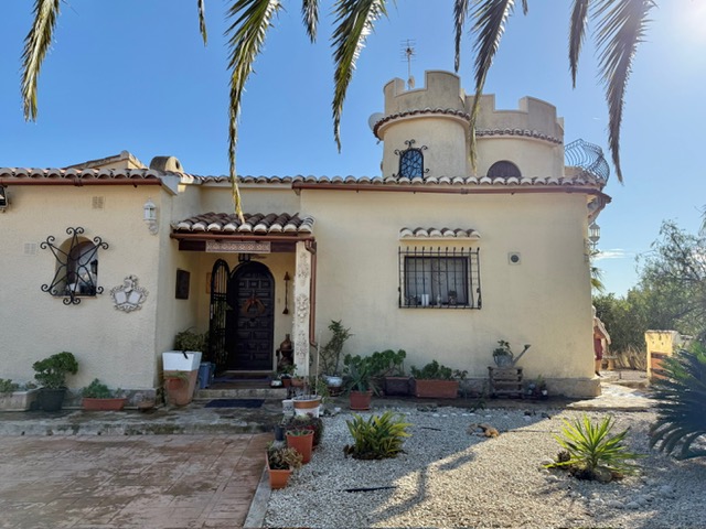 Hoofdgevel van de villa met een karakteristieke toren met kantelen, een houten boogdeur en smeedijzeren raamroosters, omgeven door een mediterrane tuin.