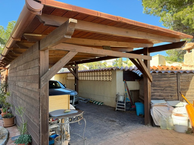 Overdekte houten carport met robuuste balken en een pannendak, voorzien van een oprit in geprint beton voor het parkeren van voertuigen.