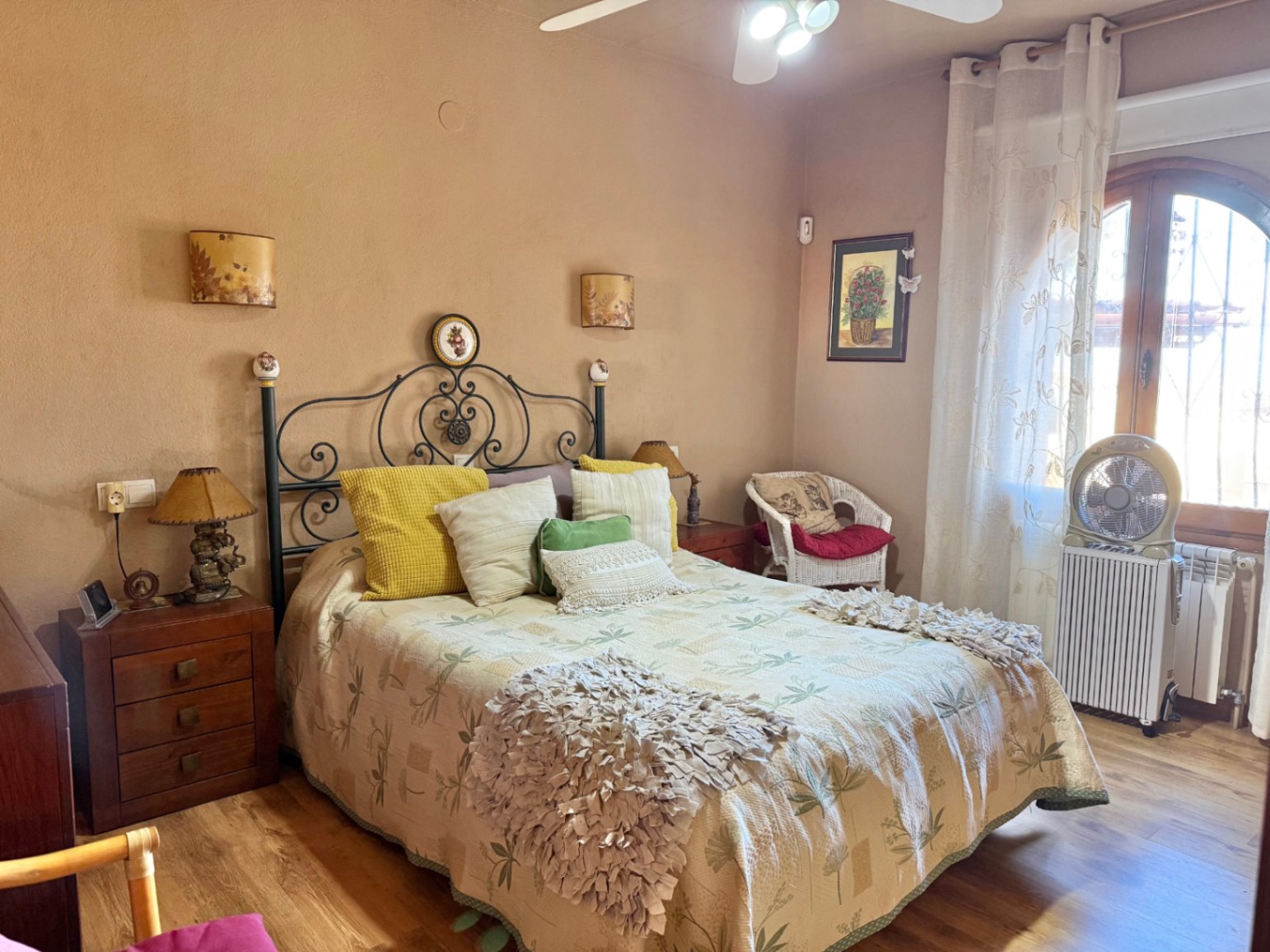 Rustieke slaapkamer met smeedijzeren hoofdbord, houten vloer en boograam. Voorzien van een plafondventilator, radiator en klassieke houten nachtkastjes.