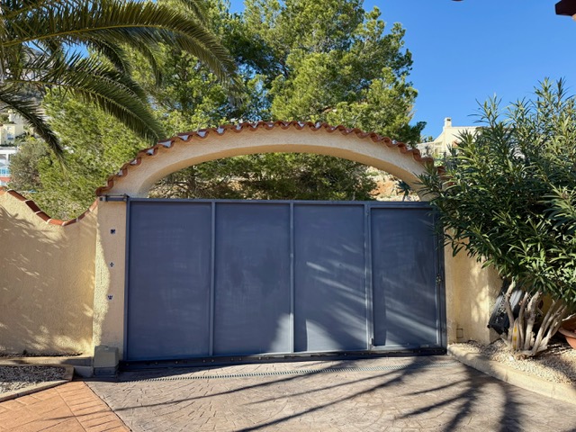 Schuifpoort onder een mediterrane boog met terracotta pannen, omgeven door volgroeide beplanting en een bestrate oprit.
