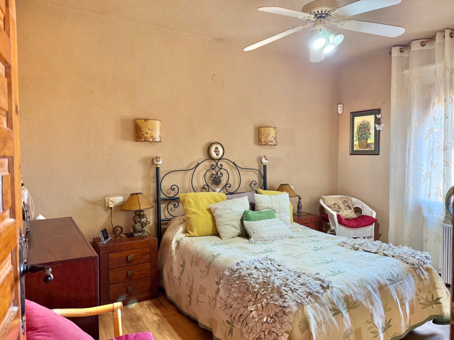 Slaapkamer met een smeedijzeren hoofdbord, houten nachtkastjes en een plafondventilator. Voorzien van een rieten fauteuil en natuurlijke lichtinval via het raam.