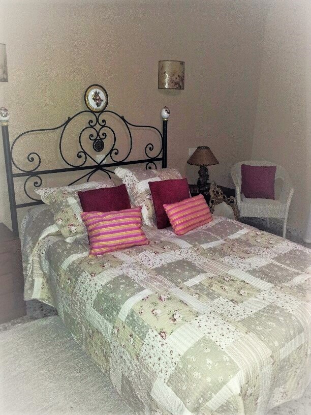 Slaapkamer met een zwart smeedijzeren hoofdbord, patchwork sprei en kleurrijke kussens. Een witte rotan stoel vult de hoek van deze traditionele kamer aan.
