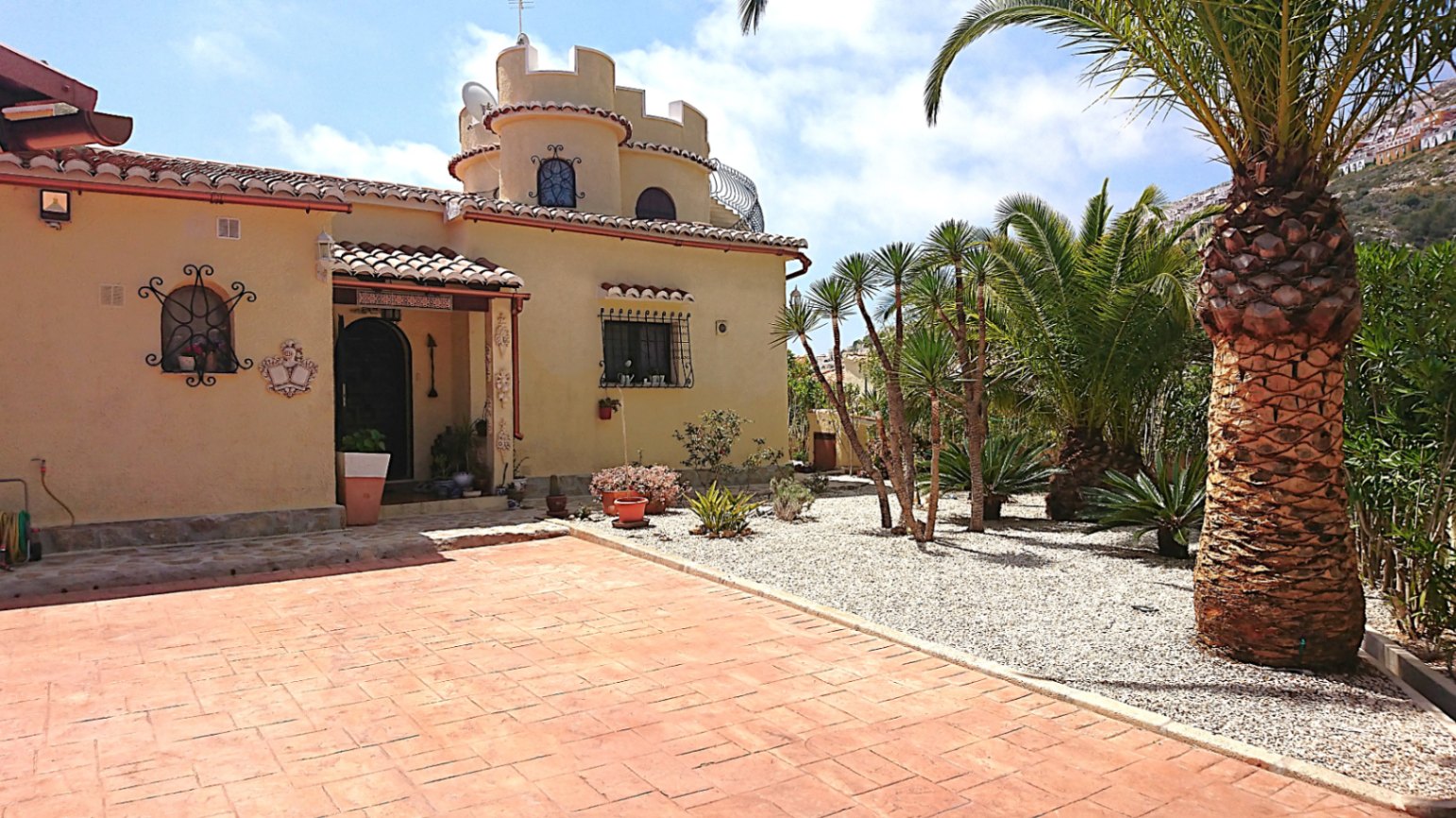 Voorgevel met een karakteristieke ronde toren, boogvensters met smeedwerk en een mediterrane tuin met volwassen palmbomen en grind.