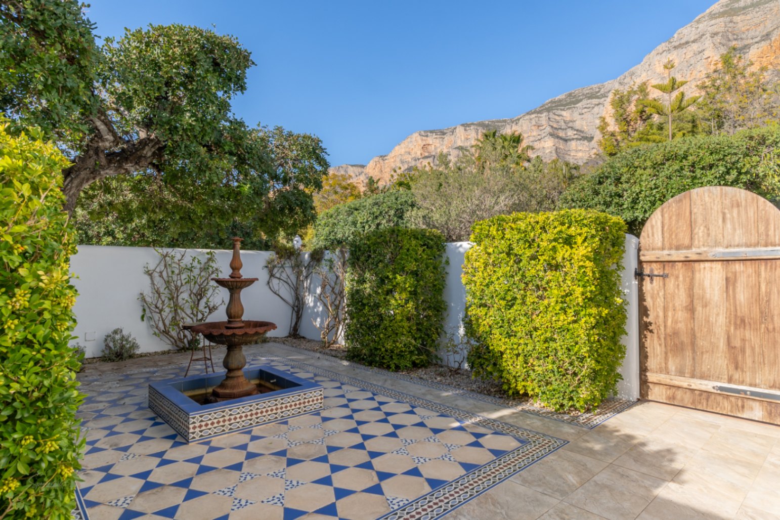 Andalusische patio met een drielaagse fontein, blauw-witte patroontegels en een rustieke houten poort met uitzicht op de Montgó-berg.