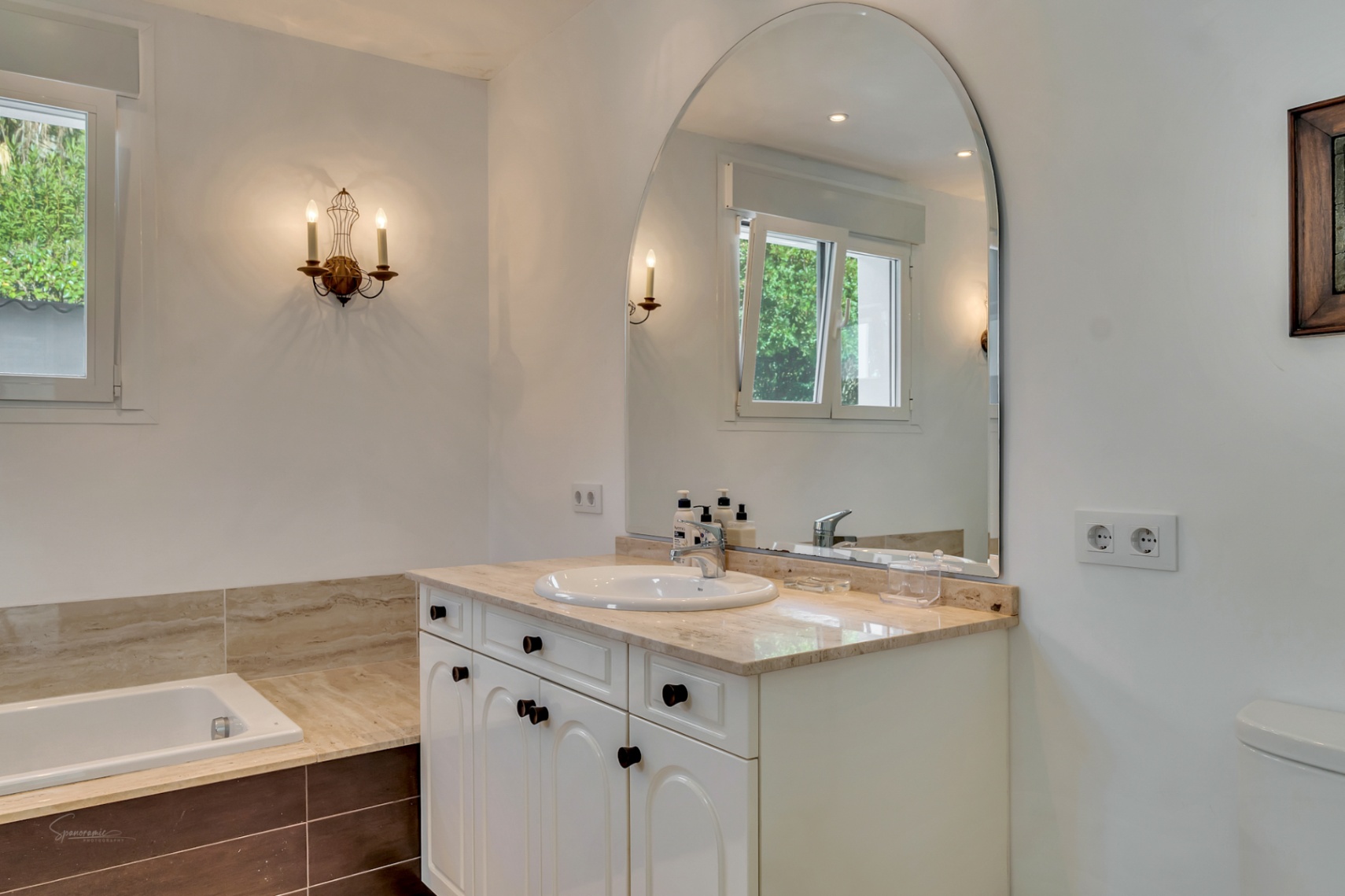 Badkamer met marmeren wastafelblad, grote boogspiegel en klassieke wandlampen. De ruimte is voorzien van een ingebouwd bad en een raam voor natuurlijke lichtinval.