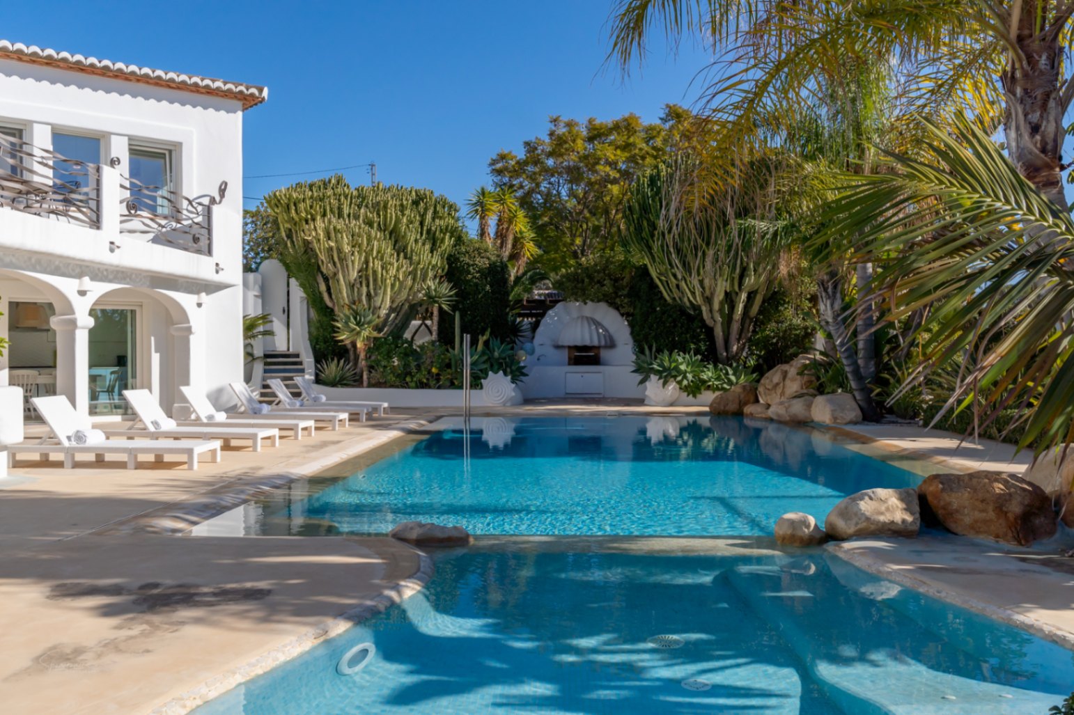 Design zwembad met hydrotherapie-gedeelte, omringd door witte ligbedden en een weelderige mediterrane tuin bij de witte villa in Ibiza-stijl.