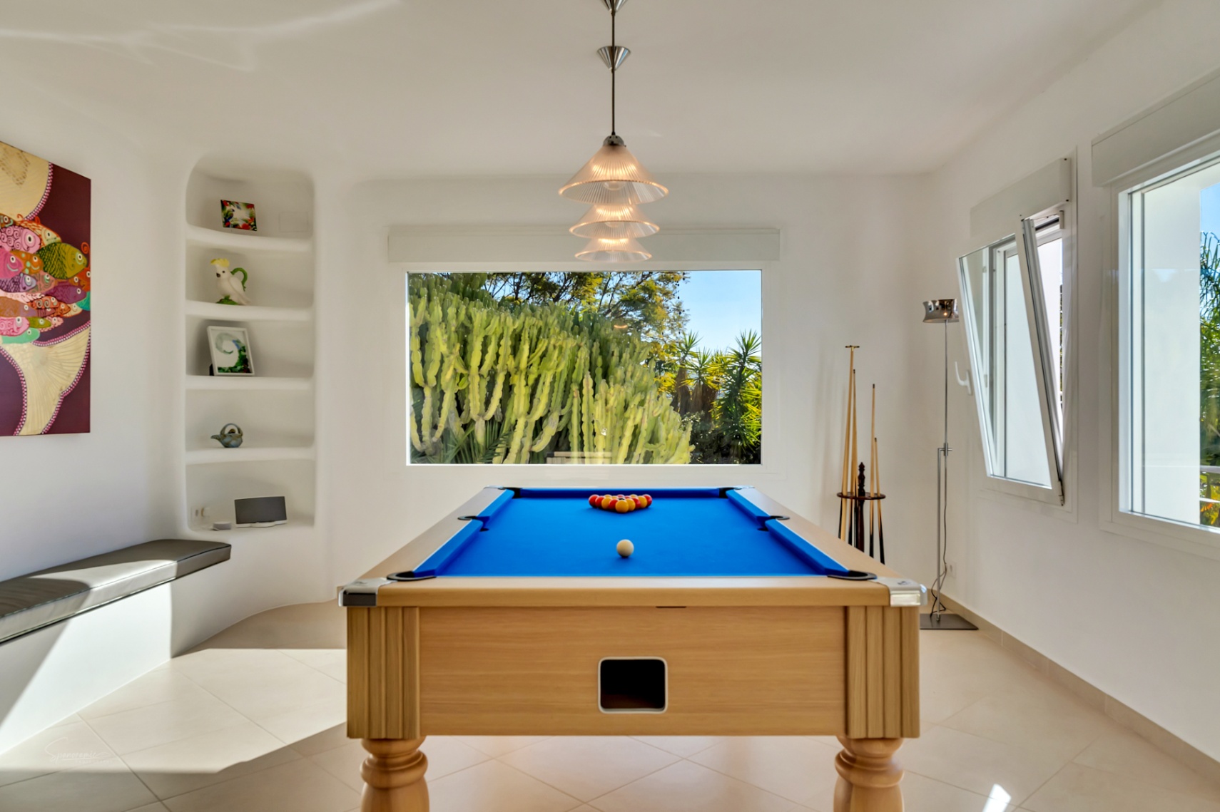 Entertainmentruimte met een houten biljarttafel met blauw laken, ingebouwde nissen en een groot raam met uitzicht op de volgroeide cactustuin.