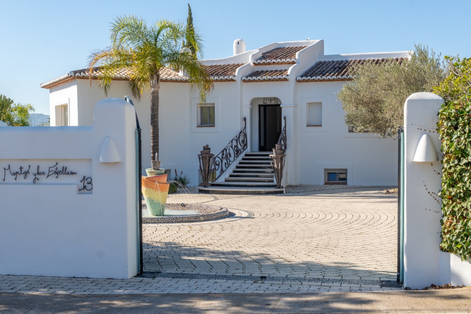Entree van een villa in Ibiza-stijl met witte gevel, een mozaïekfontein en een geplaveide binnenplaats. Sierlijke smeedijzeren leuningen flankeren de trap naar de voordeur.