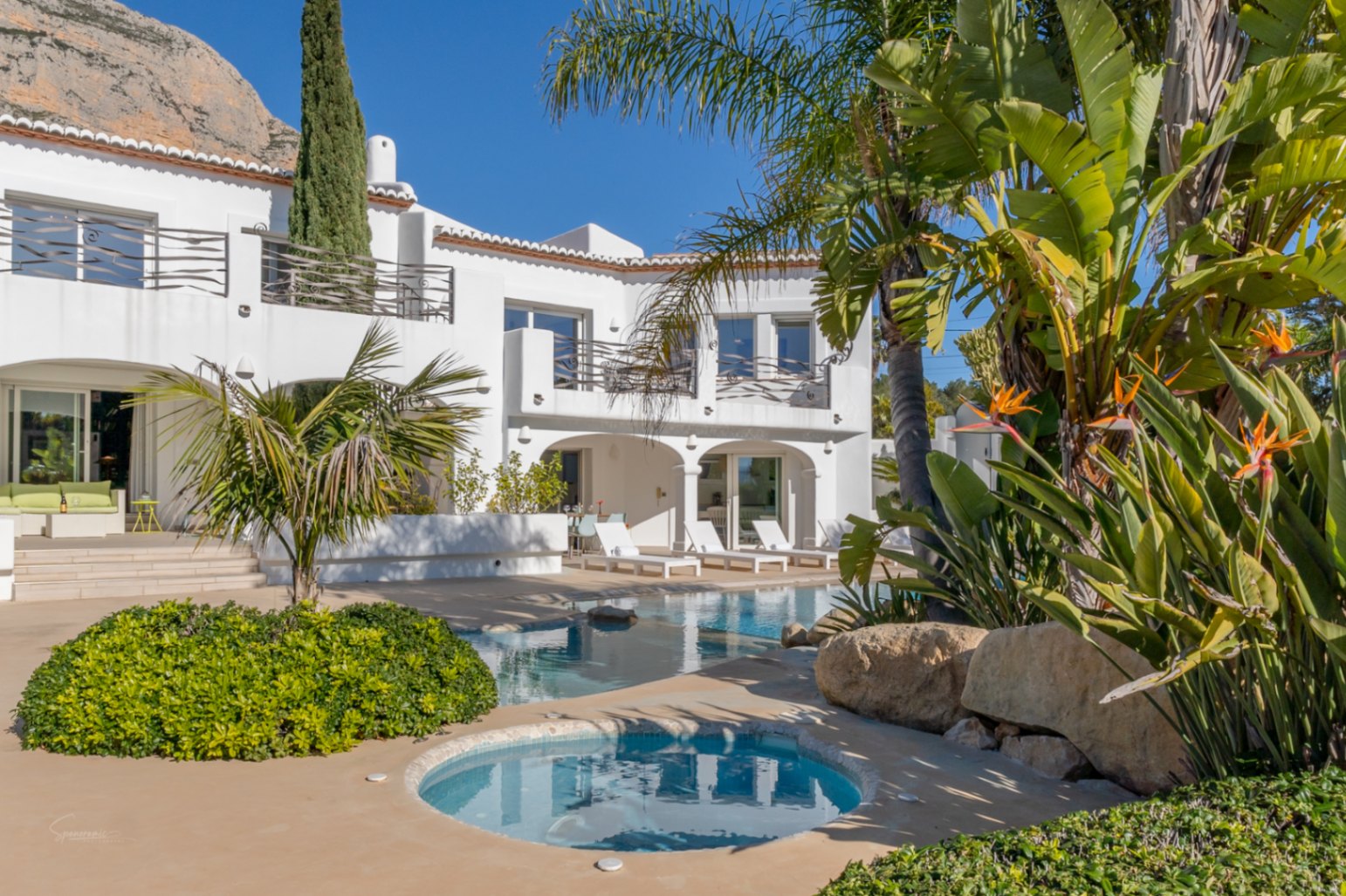 Exterieur van de villa in Ibiza-stijl met designzwembad, geïntegreerde jacuzzi en gewelfde naya tegen de achtergrond van de Montgó-berg en tropische beplanting.