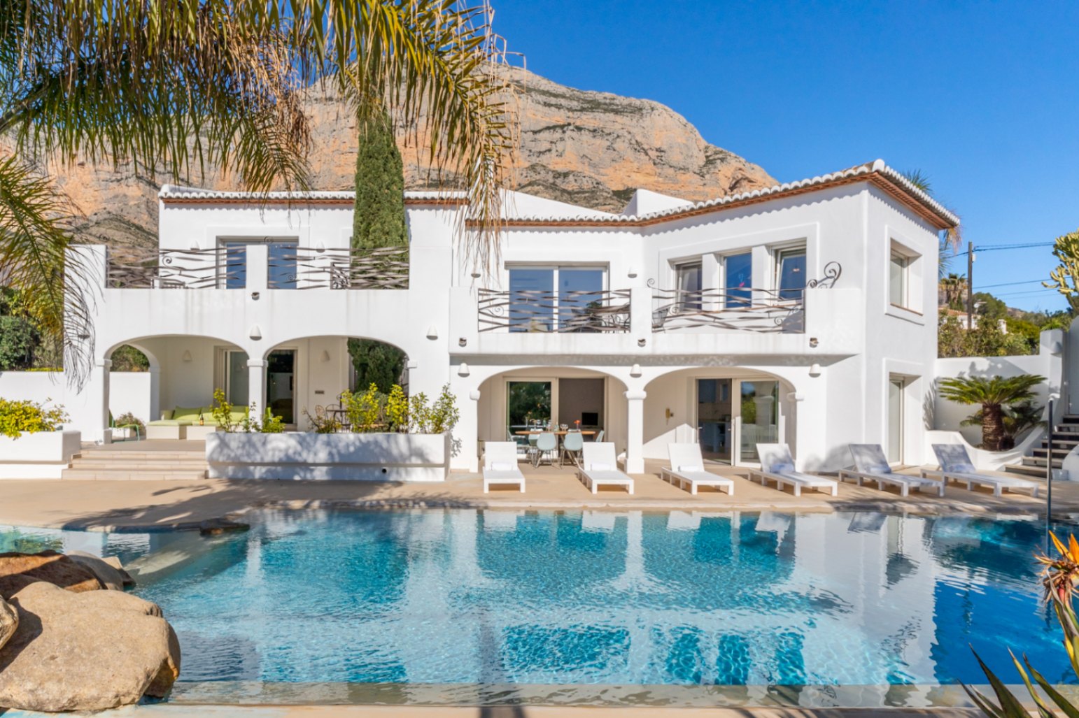 Exterieur van de villa in Ibiza-stijl met een zwembad van 70 m², overdekte terrassen met bogen en smeedijzeren hekwerk tegen de achtergrond van de Montgó.