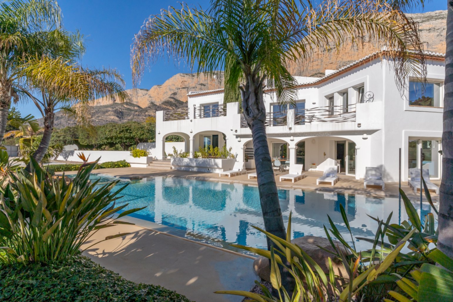 Exterieur van de villa in Ibiza-stijl met groot zwembad, overdekt terras met bogen en tropische beplanting tegen de achtergrond van de Montgó.