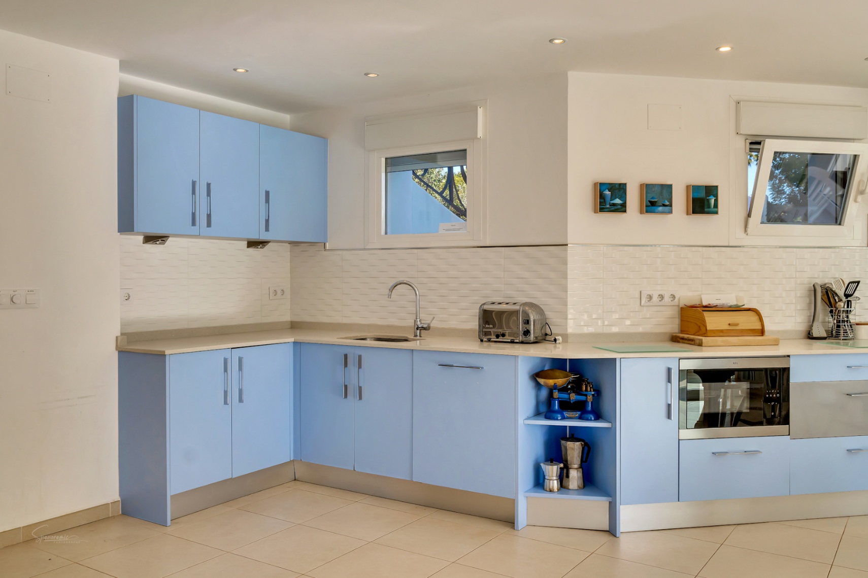Functionele keuken met pastelblauwe kasten, licht stenen werkblad en witte tegelwand. Voorzien van inbouwapparatuur en ramen voor natuurlijk lichtinval.