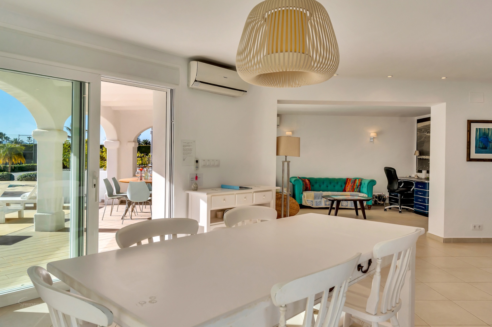 Lichte eetkamer met witte houten tafel en rieten hanglamp, verbonden met een zithoek met turquoise bank, kantoorruimte en directe toegang tot het terras.
