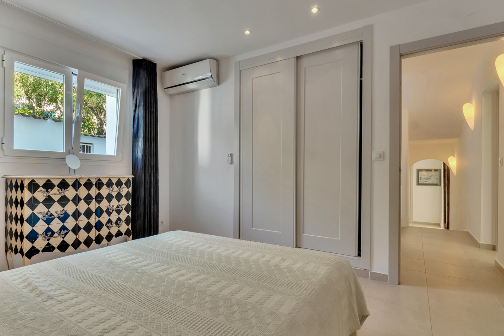 Lichte slaapkamer met inbouwkasten, airconditioning en een decoratieve ladekast. De ruimte geeft toegang tot een gang met elegante wandverlichting.