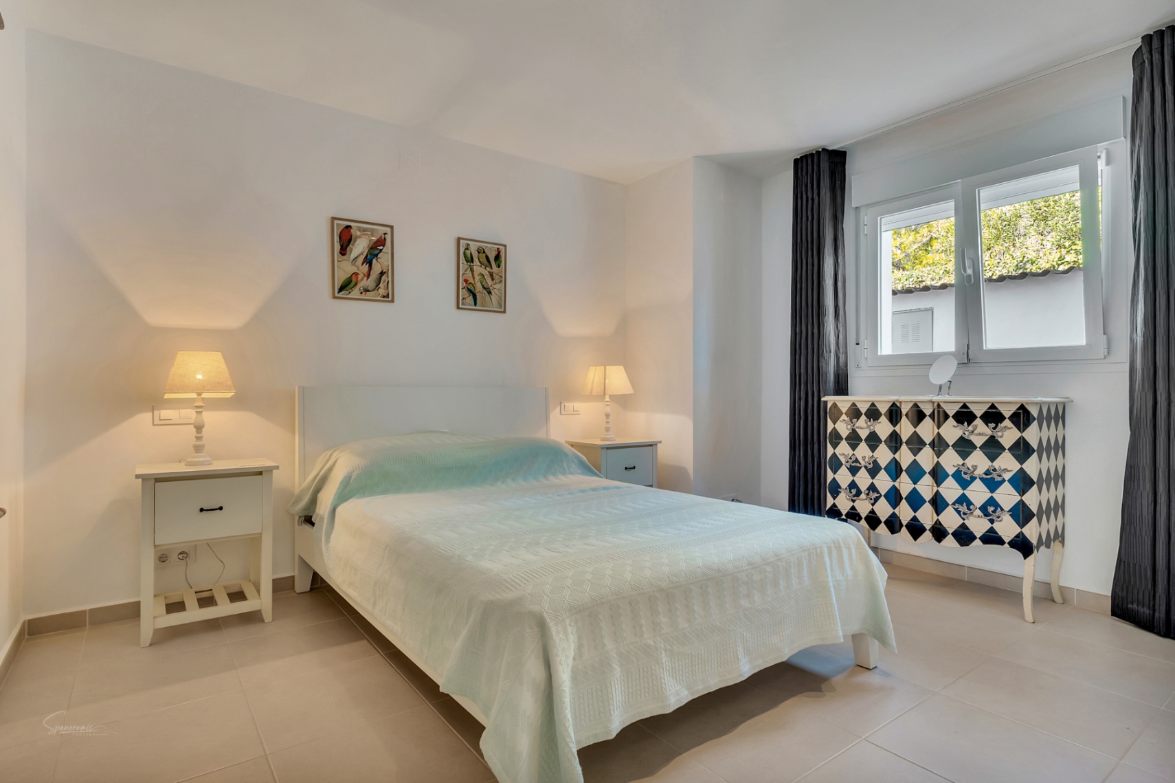 Lichte slaapkamer met tweepersoonsbed, witte nachtkastjes en een opvallende commode met ruitpatroon. Voorzien van een lichte tegelvloer en natuurlijke lichtinval.