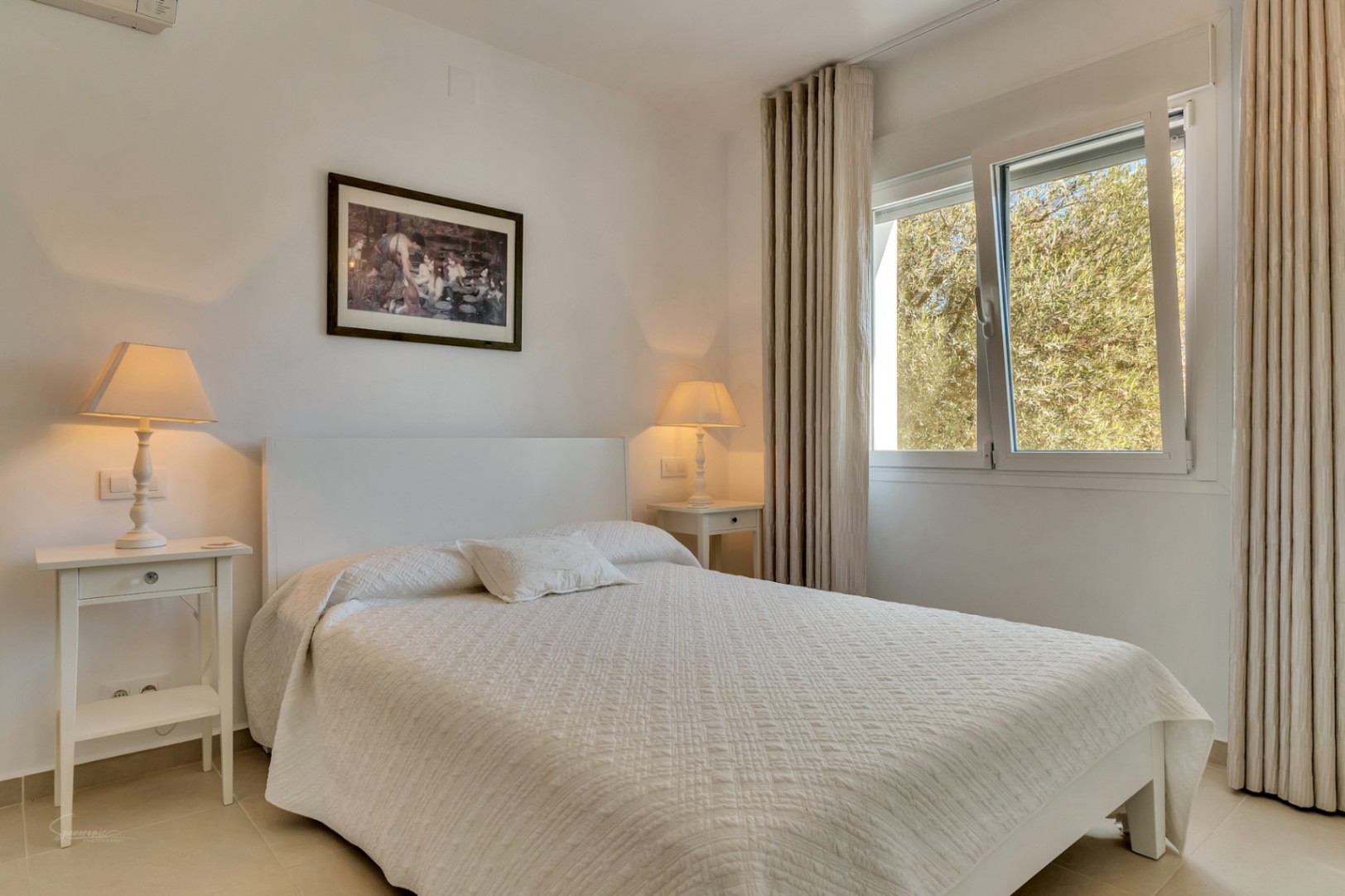 Lichte slaapkamer met wit tweepersoonsbed, bijpassende nachtkastjes en een raam met uitzicht op de tuin. Voorzien van warme verlichting en neutrale gordijnen.