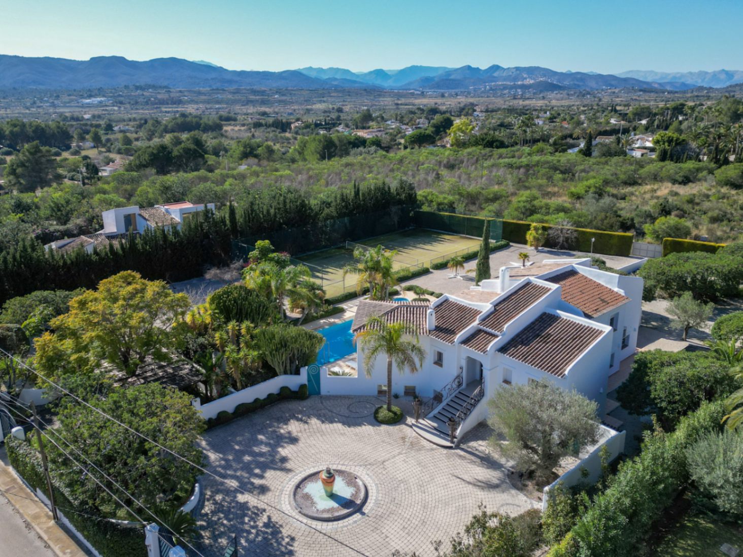 Luchtfoto van de villa in Ibiza-stijl met geplaveide oprit, centrale fontein, zwembad en tennisbaan, met uitzicht op de uitgestrekte vallei en de bergen.