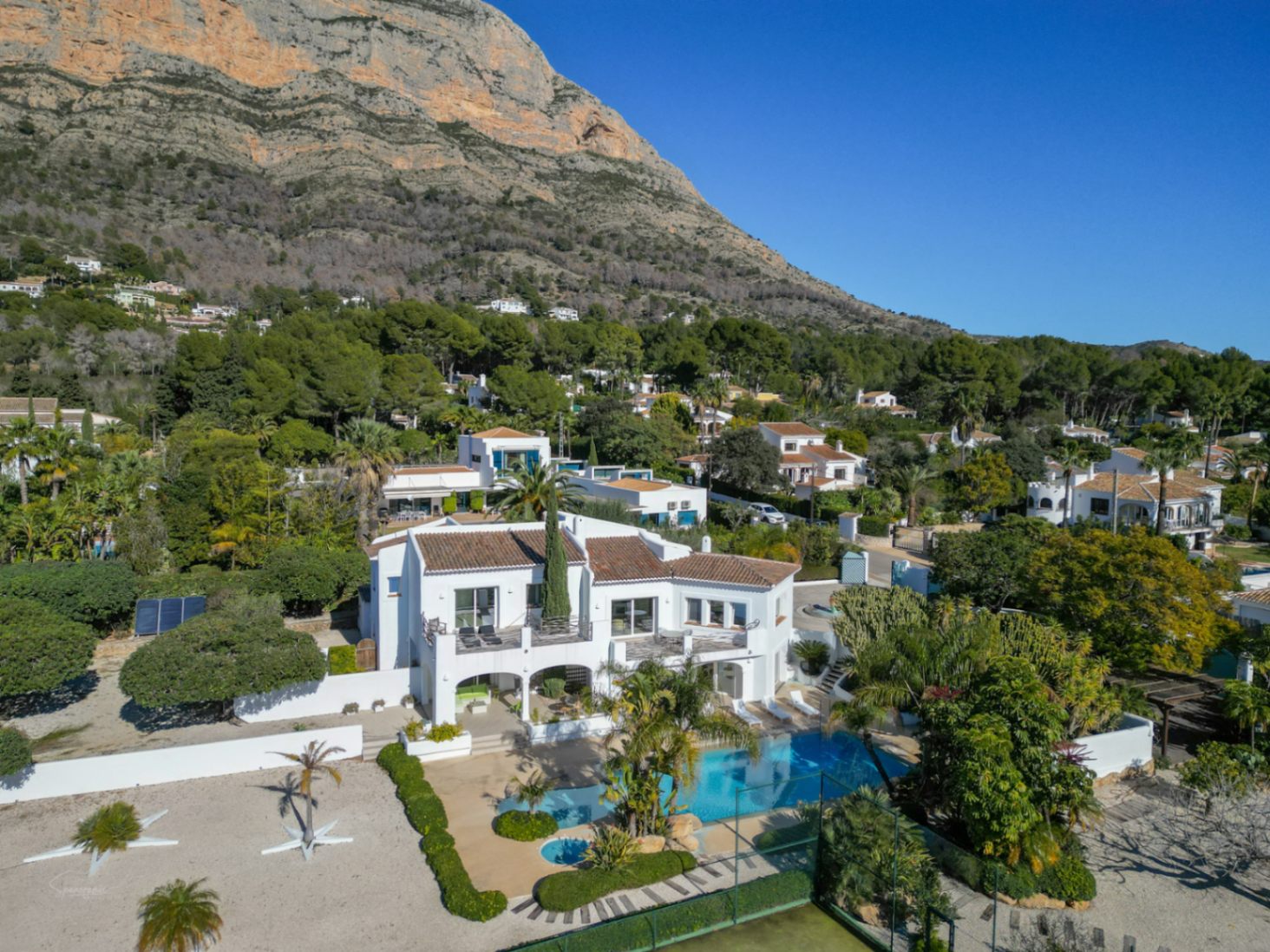 Luchtfoto van de witte villa in Ibiza-stijl met zwembad, tennisbaan en terrassen, gelegen aan de voet van de indrukwekkende Montgó-berg.